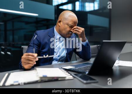 Stressed mature black businessman rubbing eyes while working laptop in office Stock Photo