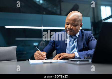 Mature black businessman writing on notepad while working with laptop in office Stock Photo