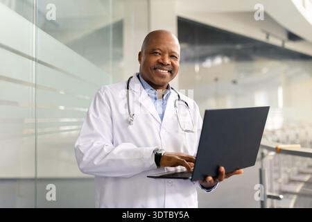 Smiling mature black doctor working on laptop in bright hospital atrium Stock Photo