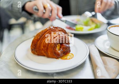 Fresh glazed croissant close up on cafe table with blurred diner and lunch plate behind, modern bakery breakfast concept with copy space and warm tone Stock Photo