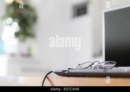 Classic reading glasses with thin black frames rest on the metallic surface of a closed laptop computer. Minimalist technology concept for digital nom Stock Photo