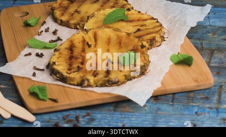Slices of caramelized grilled pineapple are garnished with whole cloves and fresh mint leaves on parchment paper. The rustic scene features a weathere Stock Photo
