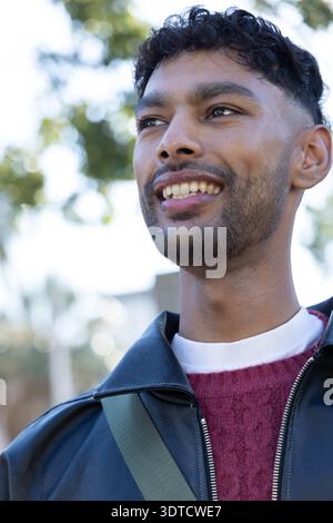 Indian man smiling, wearing black leather jacket and maroon sweater with green strap in park Stock Photo