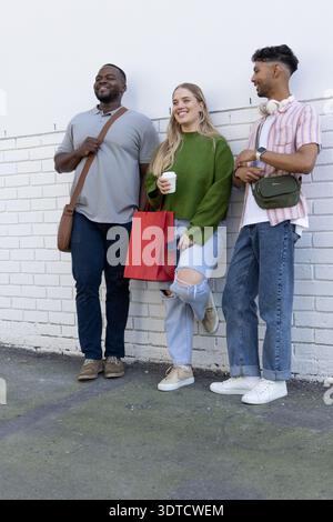 Diverse friends leaning against painted white brick wall on sidewalk, holding coffee, phone, bags Stock Photo