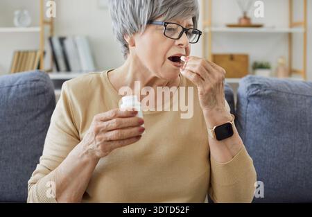 Senior woman taking pill dose for disease therapy, sitting on sofa at home Stock Photo
