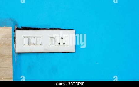 A white electrical switchboard with multiple switches and a socket installed on a bright blue wall. Stock Photo