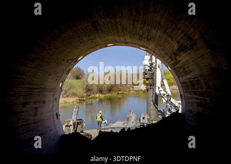 Perspective view trough pipe, from behind on worker with safety vest and white helmet managing tower drilling machine, big equipment to drill into the Stock Photo
