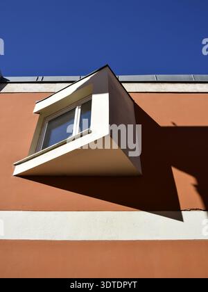 A protruding white-framed window casting a sharp angular shadow on a terracotta and beige striped wall. Stock Photo