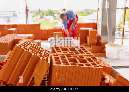 Close up shot on heap of red blocks, in background mason is building a wall with red ceramic blocks using mortar from a plastic trough. Building is un Stock Photo