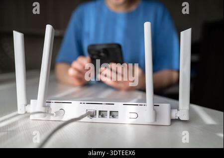 Close-up of a modern wireless router with connected cables and a woman using a smartphone in the background. Concept of home network setup, internet c Stock Photo