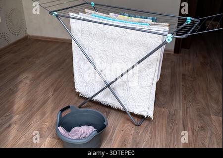 Clean laundry hanging on a grey drying rack with a laundry basket on the floor in a bright apartment. Concept of housework, laundry day, domestic rout Stock Photo