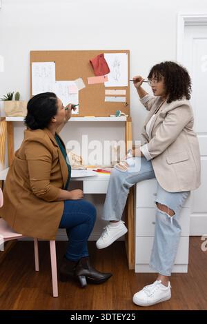 Two female designers in blazers, jeans pointing at cork board with sketches at studio holding pens Stock Photo