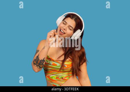 Woman with closes eyes enjoying music, wearing white headphones and singing on blue background Stock Photo