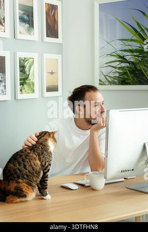 Smiling man playing with cat while working on computer from home office during coronavirus pandemic lockdown. Freelance programmer or website developer read email striking lovely pet sitting at desk || Model released Stock Photo