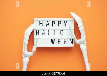 The inscription HAPPY HALLOWEEN on a lightbox on an orange background in the hands of a skeleton. Greeting card for the autumn holiday. The concept of minimalism. Eat, drink, and be scary Stock Photo