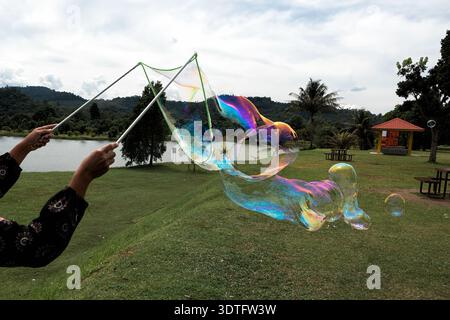 Unidentified woman Creating Giant Soap Bubbles in a lakeside park - A woman wearing a face mask uses bubble wands to create large, colorful soap bubbl Stock Photo