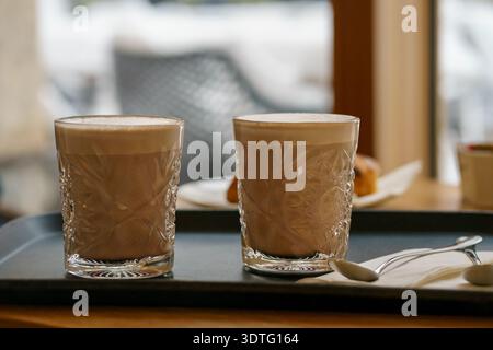 Fresh hot cocoa in clear textured glasses with thick milk foam, placed on serving tray. Warm indoor cafe setting with soft background. Stock Photo
