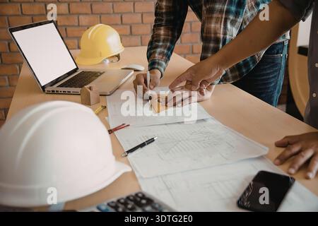 Two construction workers reviewing building plans and discussing a project on a wooden table with hard hats and a laptop. Stock Photo