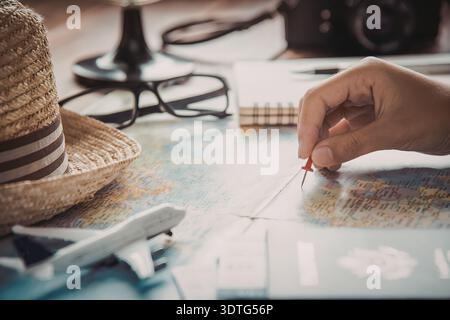 A person's hand holding a red pen pointing to a location on a map, with a straw hat, airplane model, and camera in the background. Stock Photo