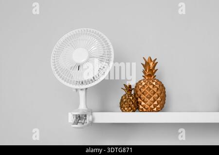 Shelf with electric fan and decorative pineapples on light wall Stock Photo