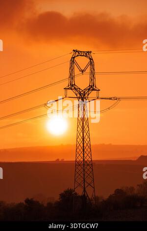 A towering electricity pylon silhouette stands majestically against a blazing orange sky at sunset. Power lines stretch across a scenic, rural landsca Stock Photo
