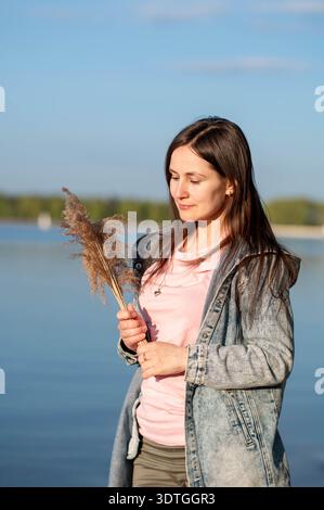 Young woman enjoying golden sunlight by the lakeside, holding rustic dried flowers in her hands Stock Photo