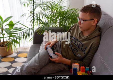 Focused boy with glasses in hoodie playing video game with wireless controller lying on grey sofa with toys and green houseplants in bright home inter Stock Photo