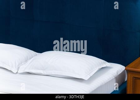 A close-up view of a plush, deep blue velvet headboard paired with neatly made white striped sheets and two soft pillows, suggesting comfort and elega Stock Photo