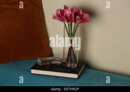 A clear vase holds pink flowers on a table, with a book open below it and glasses resting nearby, creating a simple reading area at home. Stock Photo