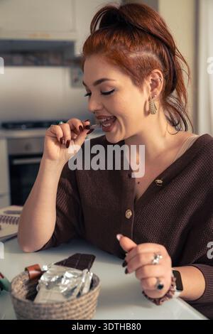 Young woman with red hair enjoying a piece of dark chocolate indoors. She is smiling while biting into the sweet treat, representing indulgence, comfo Stock Photo