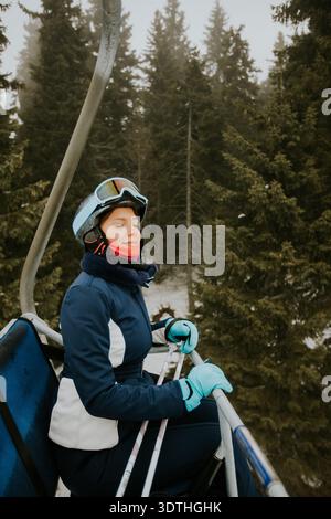 A woman sits on a ski lift wearing a ski suit and goggles. She is surrounded by tall trees covered in snow. The weather looks cloudy and cool as she e Stock Photo