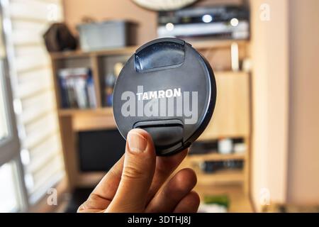 Close-up of hand holding black Tamron lens cap indoors with blurred shelving background Stock Photo