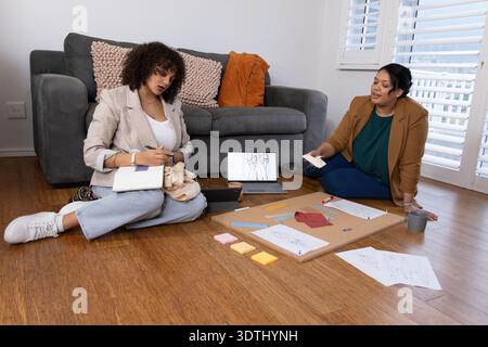 Female colleagues in blazers sitting on wood floor in living room, working over laptop and sketches Stock Photo