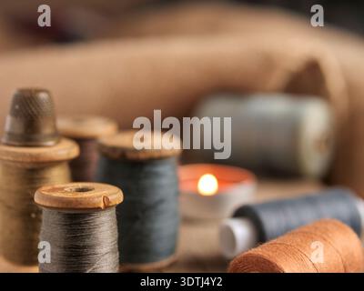 Vintage wooden thread spools by candlelight. Close-up of antique sewing notions on a rustic wooden surface with a warm glowing candle, evoking a cozy Stock Photo
