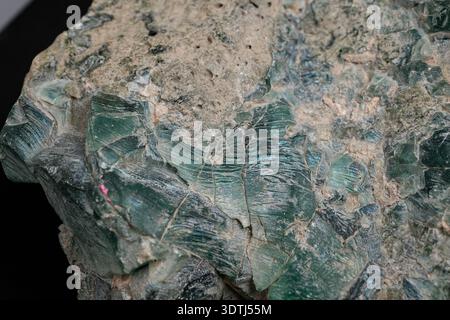 Close-up of a rough green mineral rock with layered, fibrous textures and attached beige matrix, showing cleavage lines and weathered surfaces. Stock Photo