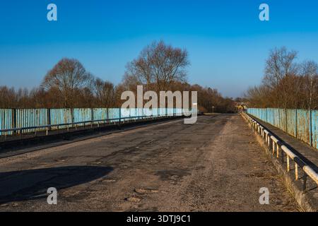 A weathered two lane bridge extends under a clear sky, with rusty pale blue railings, cracked asphalt, potholes, and distant houses on rural town outs Stock Photo