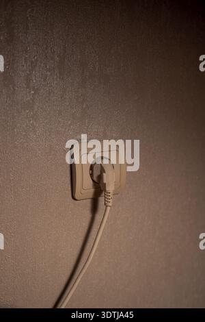 A beige plug sits in a European recessed socket on a taupe wall, indoors, under warm directional light that casts a soft cord shadow in a minimalist c Stock Photo