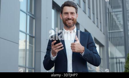 Happy Caucasian business man outdoors showing thumb up with phone smiling adult businessman using new mobile app recommend gesture looking at camera Stock Photo