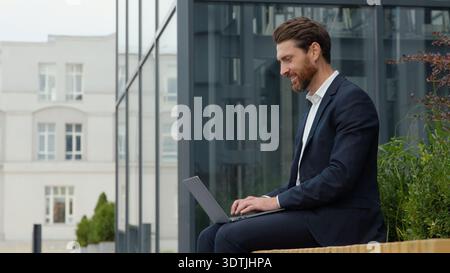 Smiling positive Caucasian mature business man typing laptop in city remote working middle-aged male professional sitting in downtown with wireless Stock Photo