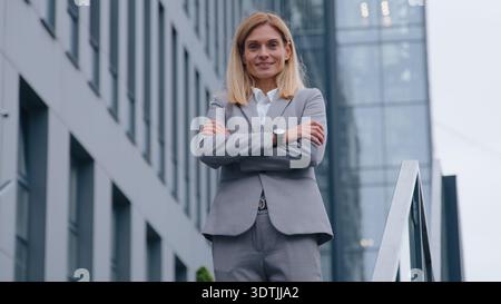 Middle-aged Caucasian business woman crossed hands posing near office building on stairs happy smiling businesswoman on stairway in downtown city Stock Photo
