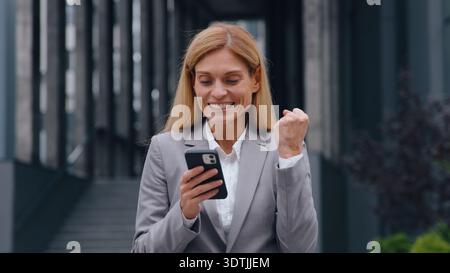 Surprised happy Caucasian adult 40s business woman with phone win in city amazed winner businesswoman winning with mobile app yes gesture female Stock Photo