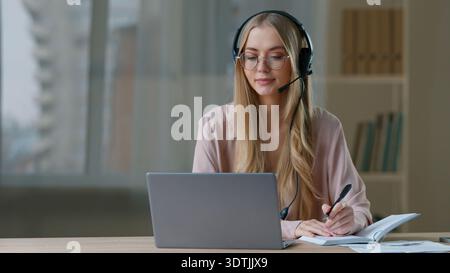 Caucasian business woman talking video call writing notes wearing headset in office businesswoman corporate studying write in notebook talk girl Stock Photo