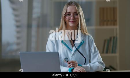 Caucasian woman female doctor posing in clinic at table medical work in hospital at desk with laptop computer girl nurse working looking at camera Stock Photo