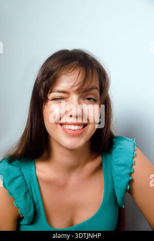 A woman smiles and winks at the camera while sitting comfortably against a simple wall in an indoor area. She shows a friendly and relaxed demeanor du Stock Photo