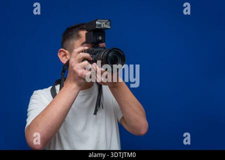 Young man holding a camera with an external flash unit, preparing to take a studio image on a blue background Stock Photo
