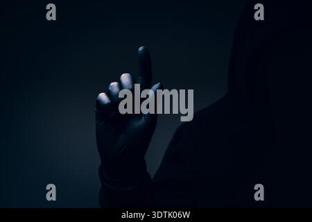 Dark hooded silhouette with obscured face and raised index finger, menacing gesture symbolizing intimidation, warning, fear, and mystery. Selective fo Stock Photo