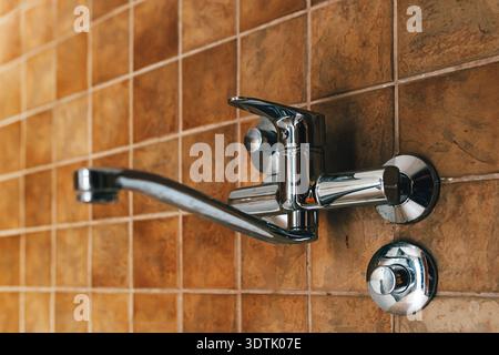 Wall-mounted chrome kitchen faucet against brown ceramic tiles, minimal interior design detail with clean reflective metal surface. Selective focus. Stock Photo