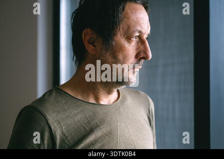 Mature adult man with unkempt hair and facial stubble looking out the window of his apartment, sad and disappointed expression, quiet reflective indoo Stock Photo