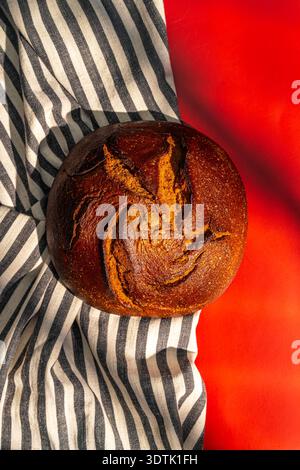 A golden-brown, freshly baked loaf of bread rests on a striped cloth, set against a vibrant red background. The artisanal crust showcases beautiful pa Stock Photo