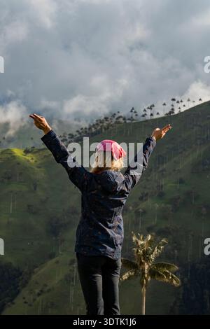 A woman joyfully raises their arms amidst the vast green landscape of the Carbonera forest in Colombia, with wax palms and misty mountains creating a Stock Photo